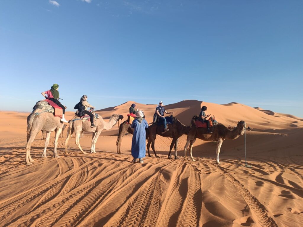 A camel guide in a blue robe with riders on camels below tall orange dunes